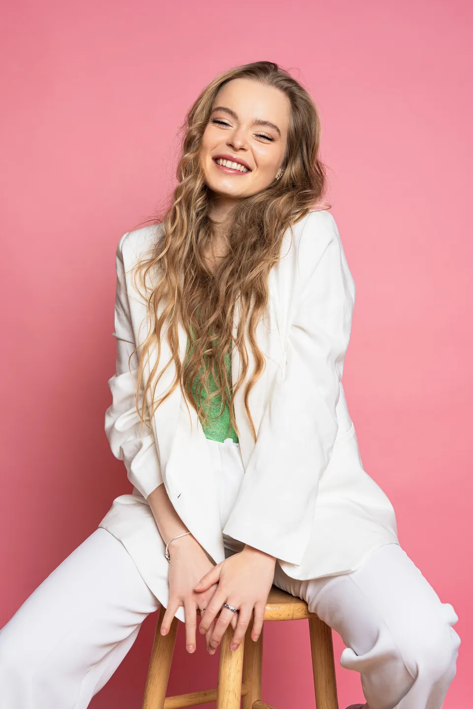 Nina Schwarz laughing, seated on a stool in a white suit against a pink backdrop