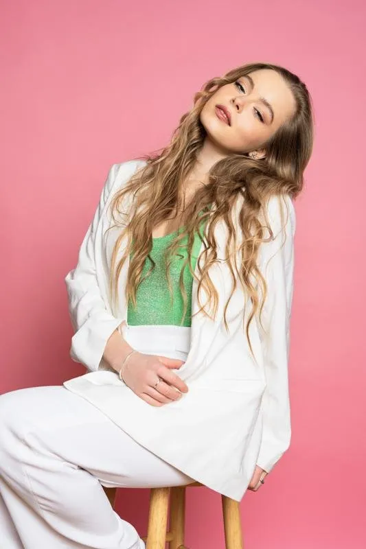 Nina Schwarz seated on a stool in a white suit, head tilted to the side